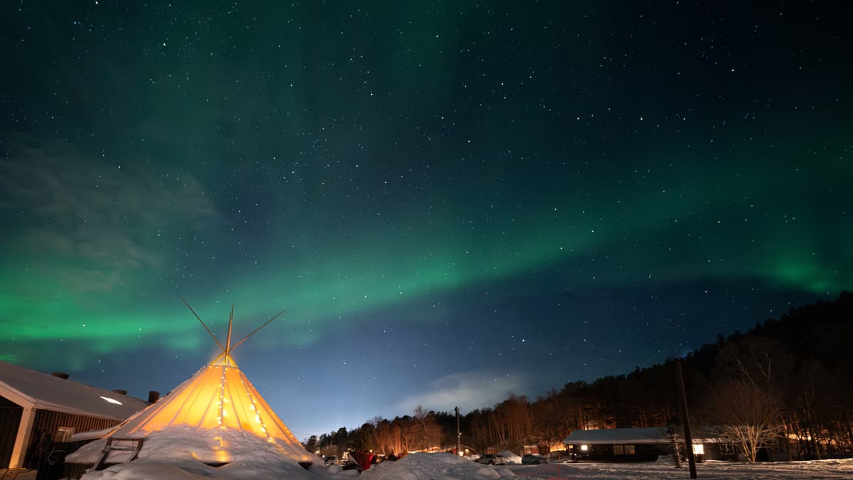 Stunning aurora borealis over a snowy landscape with a lit teepee near Alta, Norway.